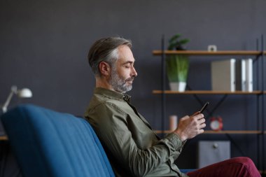 Gülümseyen gri saçlı adam cep telefonu uygulamaları kullanıyor, mesaj atıyor, internette geziniyor, internetten alışveriş yapıyor. Akıllı telefona bakmak, gülümsemek, evde oturmak. Mobil aygıtları olan olgun insanlar