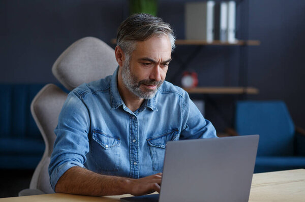 Grey-haired senior man working in home office with laptop. Business portrait of handsome manager sitting at workplace. Studying online, online courses. Business concept