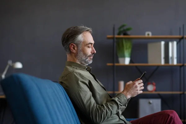 Sonriente hombre de pelo gris utilizando aplicaciones de teléfono móvil, mensaje de texto, navegación por Internet, compras en línea. Mirando el smartphone y sonriendo, sentado en casa. Personas maduras con dispositivos móviles — Foto de Stock