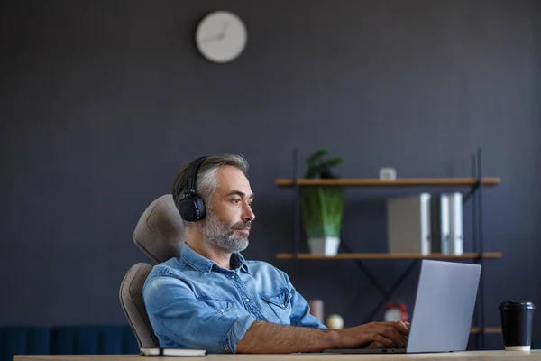 Portrait d'un bel homme aux cheveux gris portant un casque assis sur le lieu de travail. Homme freelance senior travaillant dans le bureau à domicile avec ordinateur portable. Étudier en ligne, cours en ligne — Photo