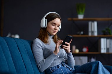 Kulaklık takan gülümseyen genç kızın portresi, mobil uygulamadan favori müzik parçalarını seçmek, mesajlaşmak, internette gezinmek, video izlemek, akıllı telefondan evde online oyun oynamak.