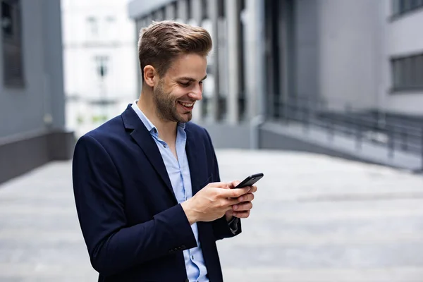 Красивый офисный работник держит смартфон и улыбается. Счастливый молодой человек использует приложения для мобильных телефонов, пишет сообщения, просматривает интернет, смотрит на смартфон. Кофе-брейк возле бизнес-центра — стоковое фото