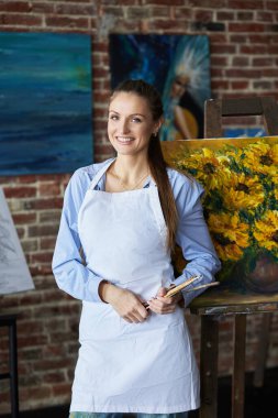 Sanat atölyesinde önlük giymiş genç, gülümseyen kadın ressamın portresi. Resim fırçası tutan bir ressam. Yaratıcı süreç, rahatlama, rahatlama, hobi, stres yönetimi