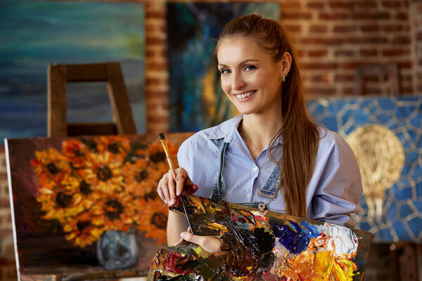 Портрет молодой улыбающейся художницы с ее произведениями в художественной студии. Художник держит кисти. Творческий процесс, отдых, досуг, хобби, стресс-менеджмент