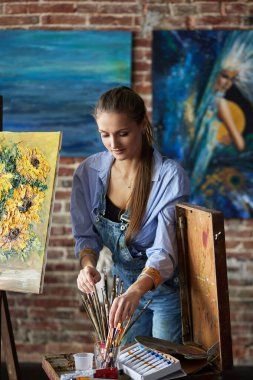 Ressam iş için fırça seçiyor. Sanat stüdyosunda gülümseyen genç bir kadın sanatçının portresi. Yaratıcı süreç, rahatlama, rahatlama, hobi, stres yönetimi.