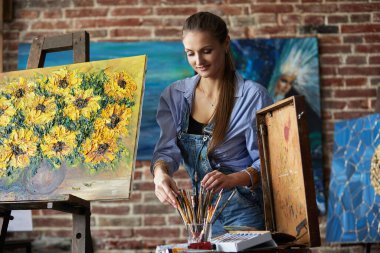 Ressam iş için fırça seçiyor. Sanat stüdyosunda gülümseyen genç bir kadın sanatçının portresi. Yaratıcı süreç, rahatlama, rahatlama, hobi, stres yönetimi.