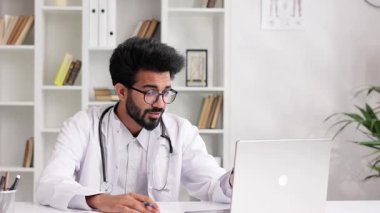Beyaz önlüklü akıllı, dost canlısı Hintli klinisyen hastane ofisinde uzaktan hasta muayenesi için dizüstü bilgisayar kullanıyor. Teletıp. Bir doktorla çevrimiçi randevu.