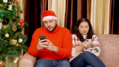 Arka planda Noel ağacı olan, koltukta oturan Hintli çift telefonu kullanıyor.