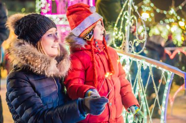 Küçük çocuk ve annesi dev köknar ağacının yanında maytap tutuyorlar ve Noel pazarında Noel aydınlatması yapıyorlar. Fuarda Noel tatili
