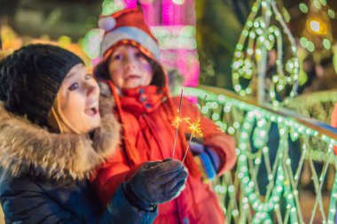 Küçük çocuk ve annesi dev köknar ağacının yanında maytap tutuyorlar ve Noel pazarında Noel aydınlatması yapıyorlar. Fuarda Noel tatili