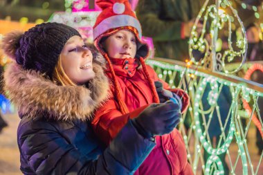 Küçük çocuk ve annesi dev köknar ağacının yanında maytap tutuyorlar ve Noel pazarında Noel aydınlatması yapıyorlar. Fuarda Noel tatili