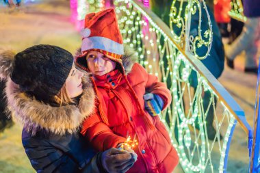 Küçük çocuk ve annesi dev köknar ağacının yanında maytap tutuyorlar ve Noel pazarında Noel aydınlatması yapıyorlar. Fuarda Noel tatili. Çılgın 2020 bitti, 2021 'deyiz.