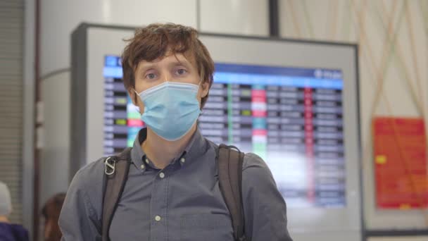 Ein junger Mann mit medizinischer Gesichtsmaske auf einem Flughafen steht in einer Halle vor einem Flugplanmonitor und blickt in die Kamera. Das Konzept der neuen Normalität des Lebensstils der Völker. Flugreisen in — Stockvideo