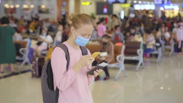 Una joven con una mascarilla médica en un aeropuerto está parada en un pasillo con un teléfono celular en sus manos. El concepto de la Nueva normalidad del estilo de vida de las personas. Viajes aéreos en la edad de Covid-19 — Vídeos de Stock