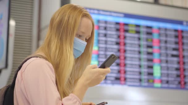 Eine junge Frau mit medizinischer Gesichtsmaske auf einem Flughafen steht mit dem Smartphone in der Hand in einer Halle vor einem Flugplanmonitor. Das Konzept der neuen Normalität des Lebensstils der Völker. Luft — Stockvideo