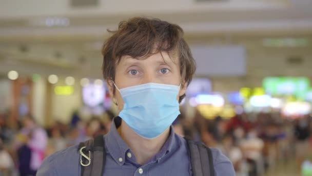 空港で医者の顔のマスクをした若い男がホールに立ってカメラを覗き込む。人々の生活様式の新しい通常の概念。コヴィト19世の時代の航空旅行 — ストック動画