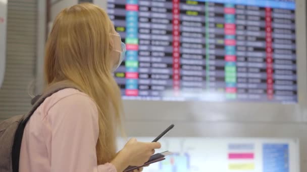 Eine junge Frau mit medizinischer Gesichtsmaske auf einem Flughafen steht mit dem Smartphone in der Hand in einer Halle vor einem Flugplanmonitor. Das Konzept der neuen Normalität des Lebensstils der Völker. Luft — Stockvideo