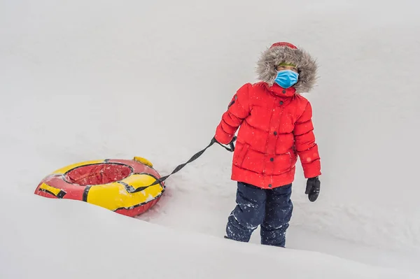 Criança se divertindo em tubo de neve usar máscaras médicas devido ao coronavírus COVID-19. O rapaz está a montar um tubo. Diversão de inverno para crianças — Fotografia de Stock