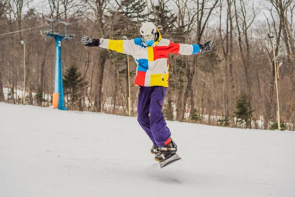 Dağlarda snowboard 'la atlayan genç adam COVID-19 Coronavirus yüzünden tıbbi maske takıyor. — Stok fotoğraf