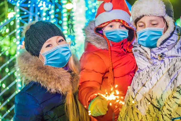 A happy family wear medical masks due to the COVID-19 coronavirus with sparklers near giant fir tree and Christmas illumination on Christmas market . Xmas holidays on fair. Crazy 2020 is over, its