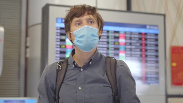 Ein junger Mann mit medizinischer Gesichtsmaske auf einem Flughafen steht in einer Halle vor einem Flugplanmonitor. Das Konzept der neuen Normalität des Lebensstils der Völker. Flugreisen im Zeitalter von Covid-19 — Stockvideo