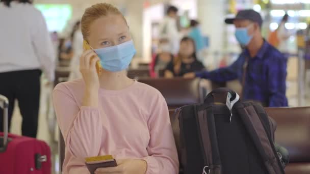 一名戴着医疗面罩的年轻女子坐在机场的椅子上，用手机交谈。人的生活方式新常态的概念。Covid-19岁时的航空旅行 — 图库视频影像