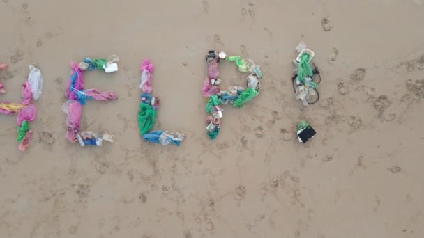 Foto aérea de una carta AYUDA hecha de basura en una playa de arena. Un triste voluntario ecológico está tirado en la arena por las letras. Detener la contaminación plástica — Vídeos de Stock
