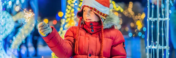 Kleiner Junge mit Wunderkerzen neben Riesentanne und Weihnachtsbeleuchtung auf dem Weihnachtsmarkt. Weihnachtsferien auf der Kirmes BANNER, LONG FORMAT — Stockfoto