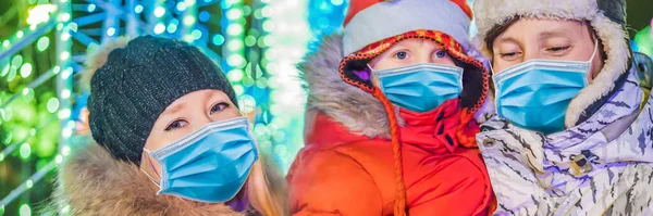 BANNER, LONG FORMAT A happy family wear medical masks due to the COVID-19 coronavirus with sparklers near giant fir tree and Christmas illumination on Christmas market . Xmas holidays on fair. Crazy