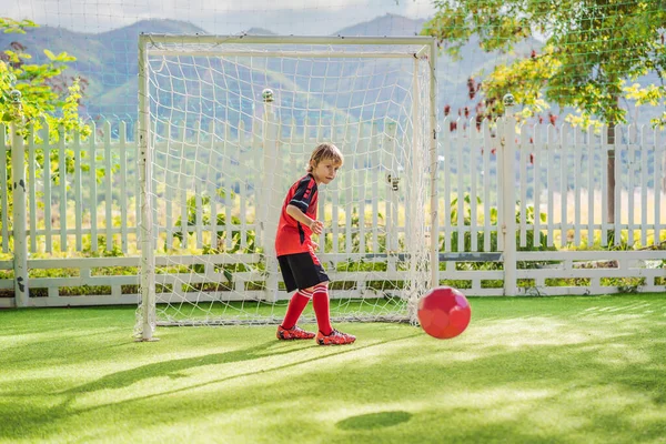 A kis cuki fiú piros foci egyenruhában focizik, focizik a pályán, a szabadban. Aktív gyermek sportolás gyerekekkel vagy apával, mosolygós boldog fiú szórakozás nyáron — Stock Fotó