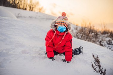 COVID-19 Coronavirus oynarken maske takan bir çocuk. Soğuk günlerde kışın çocuklarla aktif bir eğlence. Çocuk ilk karla eğleniyor. Mutlu küçük çocuk karda oynuyor, aferin.