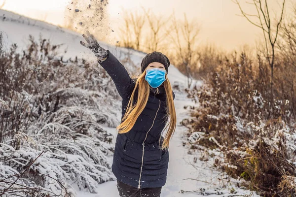 Inverno ragazza indossa una maschera medica durante COVID-19 coronavirus lancio palla di neve alla macchina fotografica sorridente felice divertirsi all'aperto sulla neve giornata invernale giocando nella neve. Carino giocoso giovane donna all'aperto — Foto Stock
