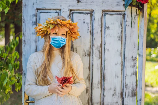 在《 COVID-19 》中，户外生活方式与戴着医疗面具、头戴秋天叶子花环的迷人金发年轻女性的肖像非常接近。穿着时髦的针织套头衫。的花环 — 图库照片