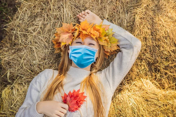 Estilo de vida al aire libre de cerca retrato de la encantadora mujer joven rubia con una máscara médica durante el coronavirus COVID-19 con una corona de hojas de otoño en un pajar. Usar un elegante jersey de punto — Foto de Stock