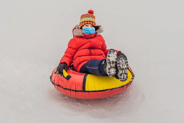 Niño usando una máscara médica durante el coronavirus COVID-19 divirtiéndose en el tubo de nieve. El chico está montando un tubo. Diversión de invierno para niños —  Fotos de Stock