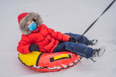 Çocuk COVID-19 Coronavirus sırasında tıbbi maske takarak kar tüpünde eğleniyor. Çocuk bir boruya biniyor. Çocuklar için kış eğlencesi