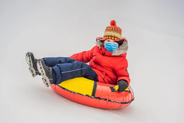 Dítě s lékařskou maskou během COVID-19 coronavirus baví na sněhu trubice. Ten kluk jezdí na trubkách. Zimní zábava pro děti — Stock fotografie