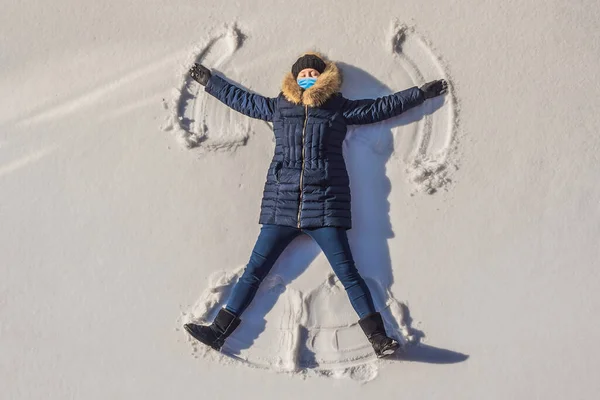 Retrato de una hermosa mujer joven con una máscara médica durante el coronavirus COVID-19 acostada en un lago de nieve congelado moviendo sus brazos y piernas hacia arriba y hacia abajo creando una figura de ángel de la nieve, jugando — Foto de Stock
