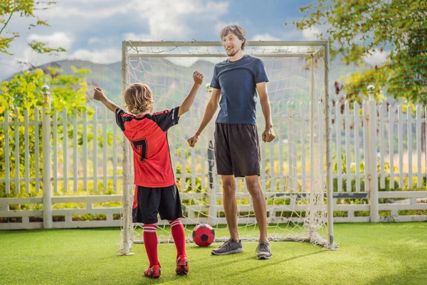 Kleiner süßer Junge in roter Fußballuniform und sein Trainer oder Vater spielen Fußball, Fußball auf dem Feld, im Freien. Aktives Kind, das mit Kindern oder Vater Sport treibt, lächelnder fröhlicher Junge, der Spaß an — Stockfoto