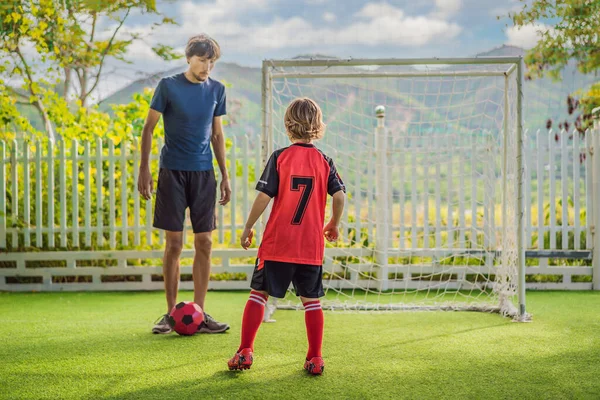 Pequeno menino bonito em uniforme de futebol vermelho e seu treinador ou pai jogando futebol, futebol no campo, ao ar livre. Criança ativa fazendo esportes com crianças ou pai, sorrindo menino feliz se divertindo em — Fotografia de Stock