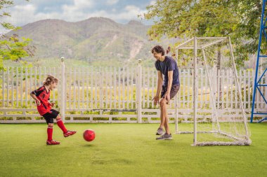 Kırmızı futbol forması içinde küçük şirin bir çocuk ve antrenörü ya da babası futbol oynuyor, sahada, dışarıda. Aktif çocuk çocuklarla ya da babayla spor yapıyor, gülümseyen mutlu çocuk eğleniyor.