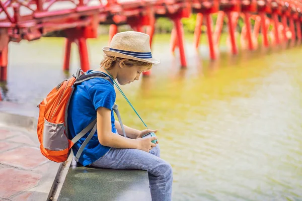 Vit pojke turist på bakgrund av Red Bridge i offentlig park trädgård med träd och reflektion i mitten av Hoan Kiem Lake i centrala Hanoi. Resa med barn koncept — Stockfoto