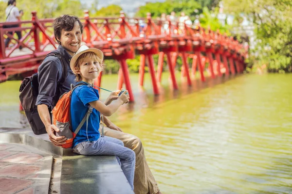 Kavkazský táta a syn cestovatelé na pozadí Red Bridge v zahradě veřejného parku se stromy a odraz uprostřed jezera Hoan Kiem v centru Hanoje. Koncept cestování s dětmi — Stock fotografie