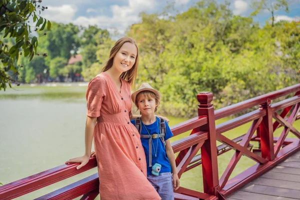 ハノイ中心街のホアンキエム湖の真ん中に木や反射と公共公園の庭でレッドブリッジを背景に白人の母親と息子の旅行者。子供と一緒に旅行するコンセプト — ストック写真