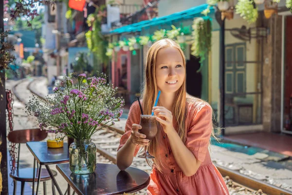 Mladá žena cestovatel pije kávu sedí u železničních drah, které procházejí obytné čtvrti v Hanoji města. Hanoi Train Street je známá turistická destinace. Vietnam se znovu otevře po koronaviru — Stock fotografie