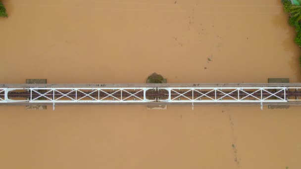 Εναέρια αργή κίνηση βολή μιας μεταλλικής σιδηροδρομικής γέφυρας πάνω από ένα ποτάμι, το οποίο έχει ένα καφετί-κίτρινο χρώμα — Αρχείο Βίντεο