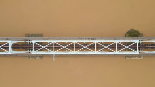 Εναέρια αργή κίνηση βολή μιας μεταλλικής σιδηροδρομικής γέφυρας πάνω από ένα ποτάμι, το οποίο έχει ένα καφετί-κίτρινο χρώμα — Αρχείο Βίντεο