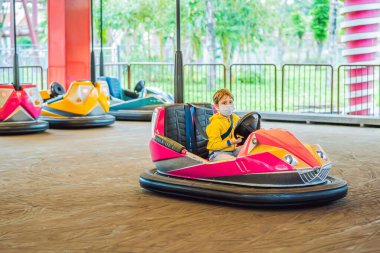 COVID-19 Coronavirus sırasında tıbbi maske takan bir çocuk lunaparktaki çarpışan arabaya biniyor.