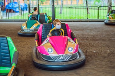 COVID-19 Coronavirus sırasında tıbbi maske takan bir çocuk lunaparktaki çarpışan arabaya biniyor.