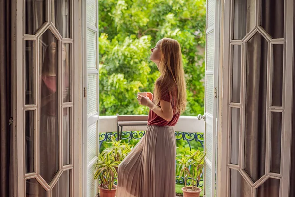 Una mujer pelirroja se encuentra en un balcón de estilo patrimonial disfrutando de su café de la mañana. Una mujer en un hotel en Europa o Asia mientras el turismo se recupera de una pandemia. El turismo se ha recuperado gracias a —  Fotos de Stock